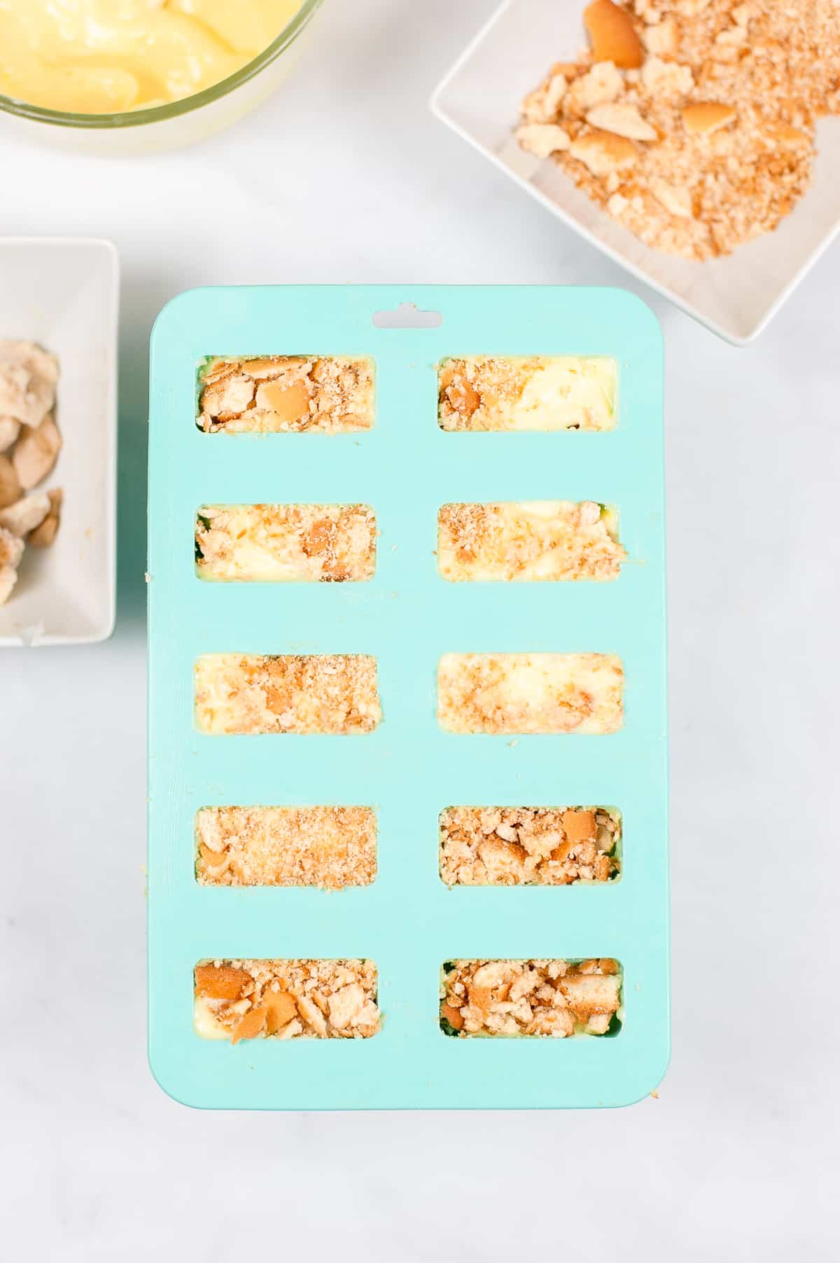 popsicle mold with cavities for 10 popsicles. Filled with banana cream pudding and vanilla wafer crumbles. More Nilla crumbs, banana slices, and pudding can be seen in background