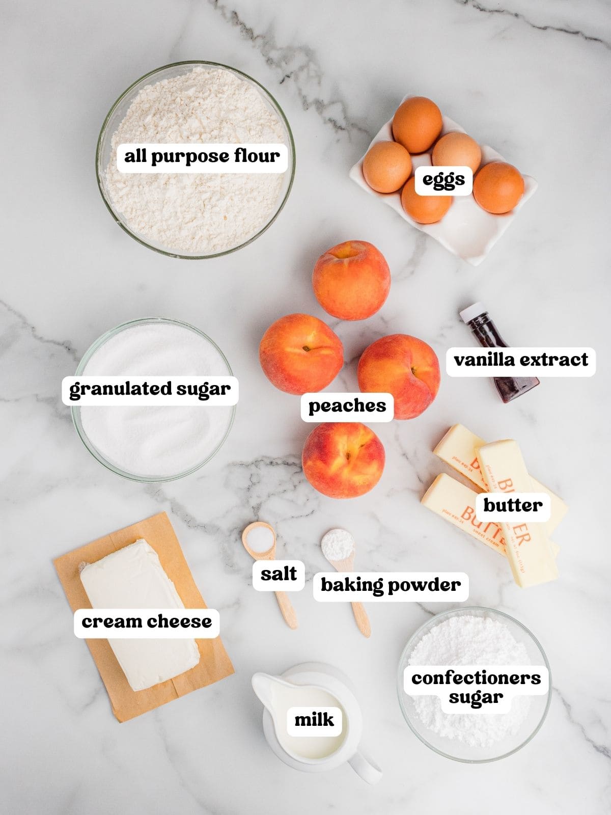 Overhead view of ingredients arranged on a marble surface for peach cake, each labeled: all purpose flour, eggs, granulated sugar, peaches, vanilla extract, butter, cream cheese, salt, baking powder, milk, and confectioners sugar.