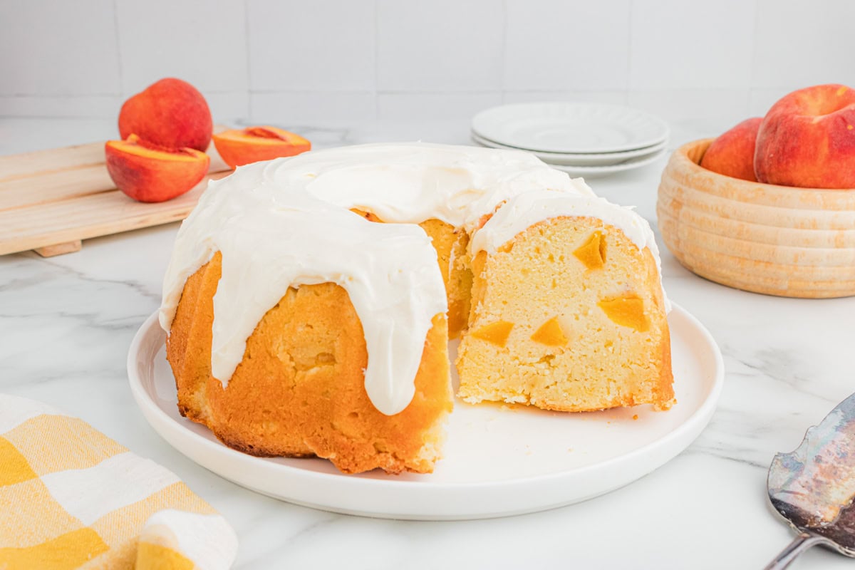 Peach bundt cake with cream cheese icing on a plate, sliced to show peach pieces inside, with fresh peaches in the background.