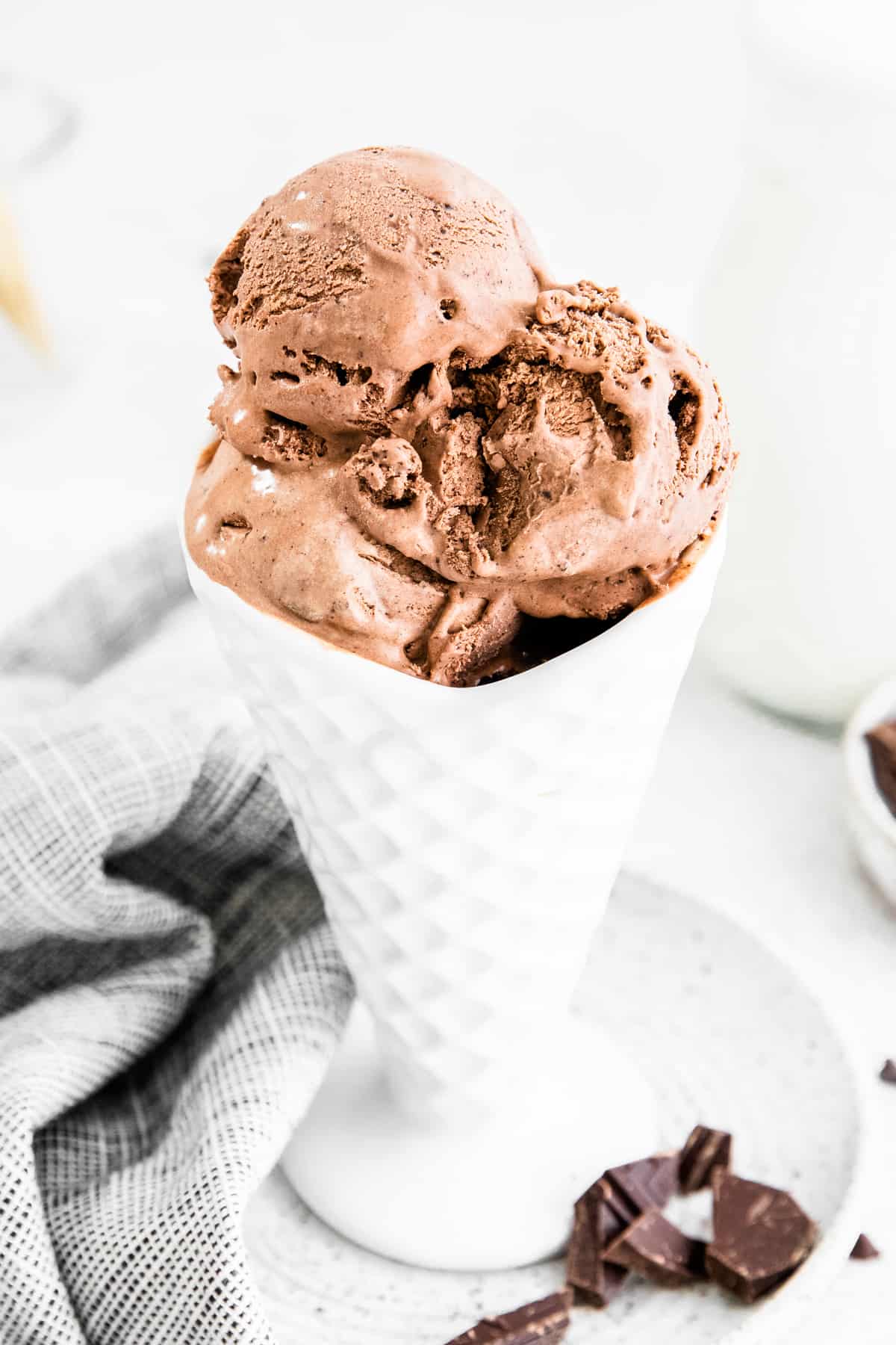 Homemade chocolate ice cream served in ice cream cone shaped bowl. Chopped chocolate and linen can be seen around the bowl as well