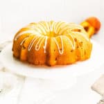 Golden bundt cake topped with drizzle of icing. Cake is on white cake stand and fresh peaches are in background.