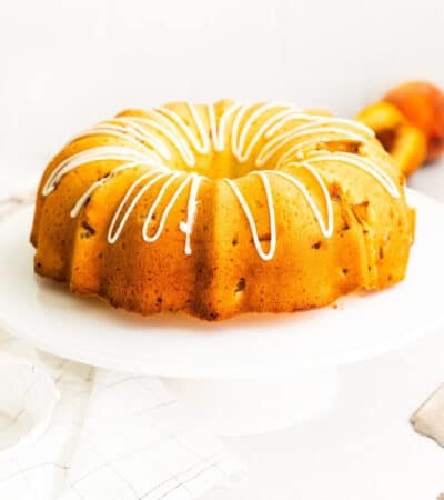 Golden bundt cake topped with drizzle of icing. Cake is on white cake stand and fresh peaches are in background.