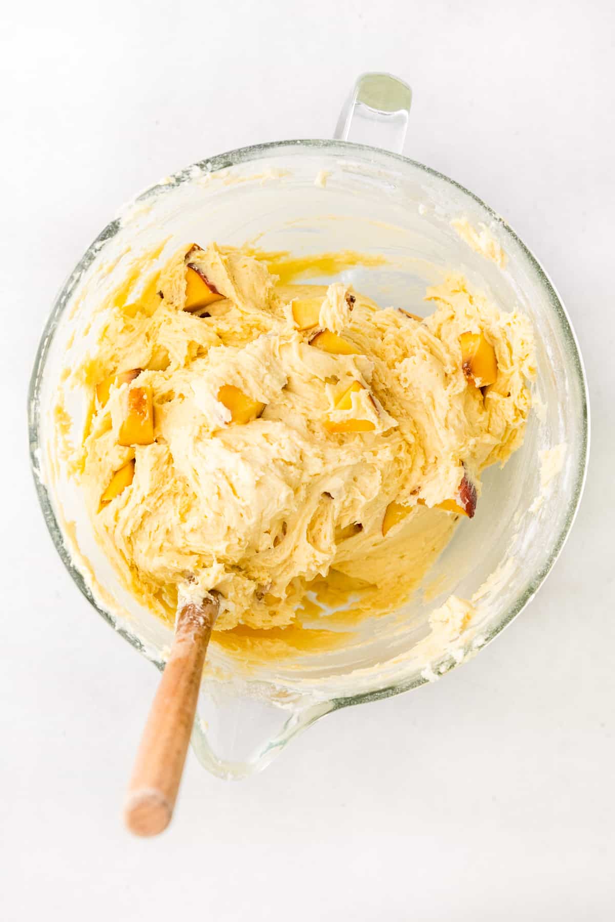Glass mixing bowl with thick peach bundt cake batter in it, sticking to a wooden spoon