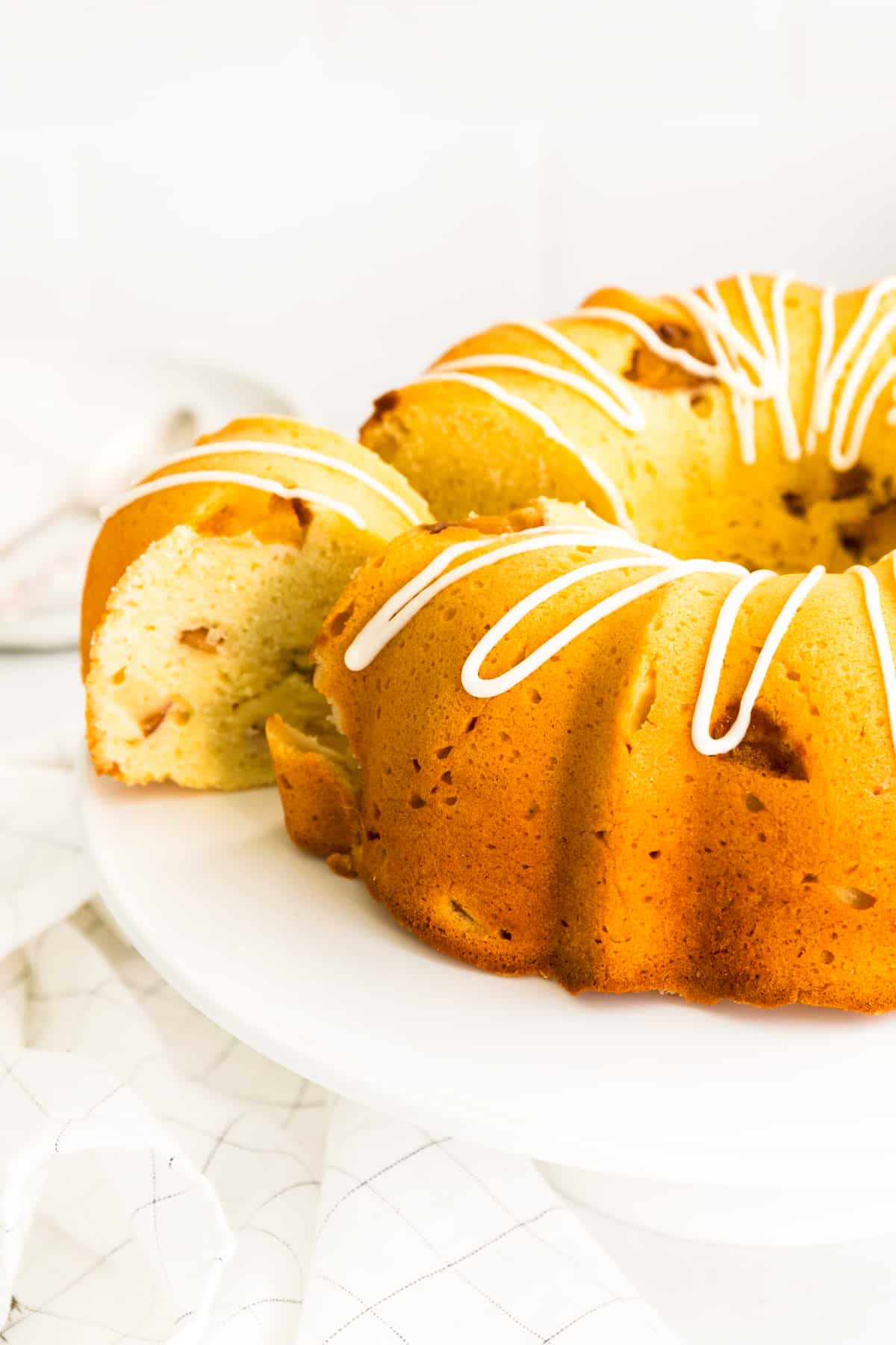 Golden brown bundt cake filled with chunks of fresh peaches and topped with drizzle of icing. A slice is being removed with cake server.