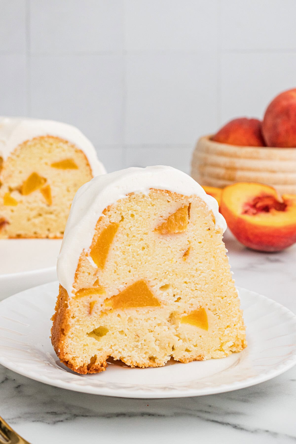 A slice of peach pound cake with cream cheese frosting on a plate with fresh peaches and rest of the cake in the background.