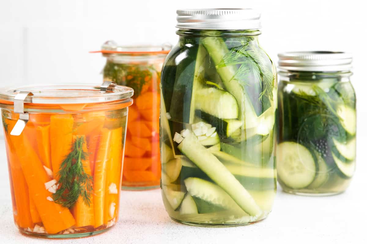 Glass jars with sliced pickled cucumbers. More jars in background, some with pickled cucumbers and others with carrots.