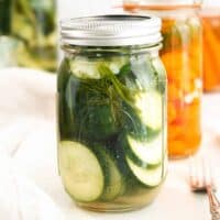 Wide Mouth Mason Jar filled with sliced pickled cucumbers. More jars in background, some with pickled cucumbers and others with carrots.