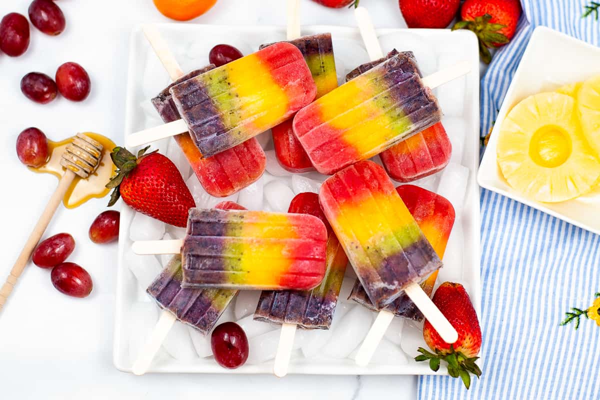 Rainbow popsicles served on white platter and surrounded by fresh fruit and a dribble or honey