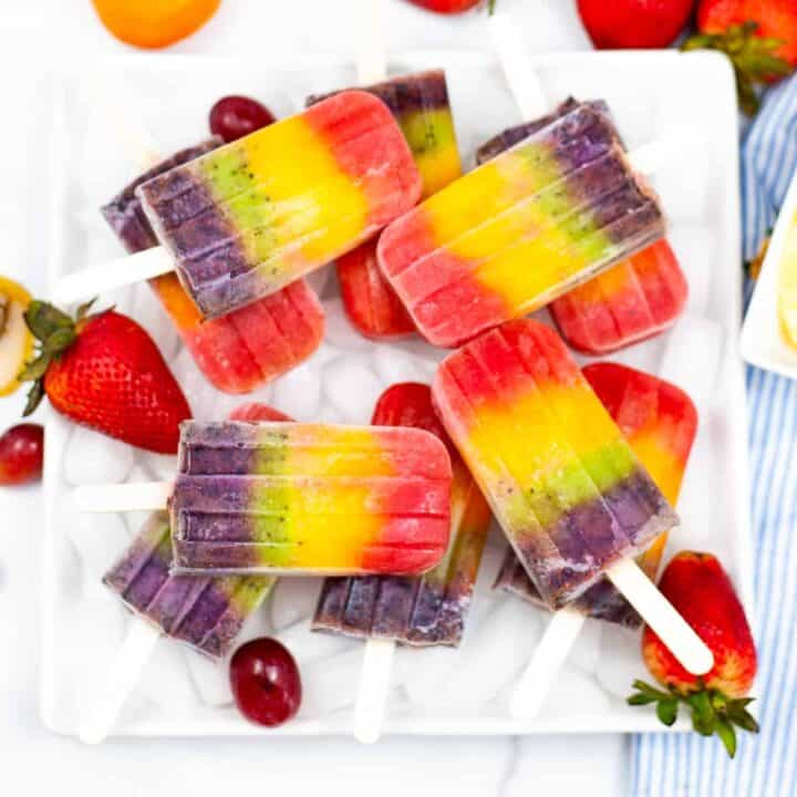 Rainbow popsicles served on a white platter garnished with strawberries and grapes