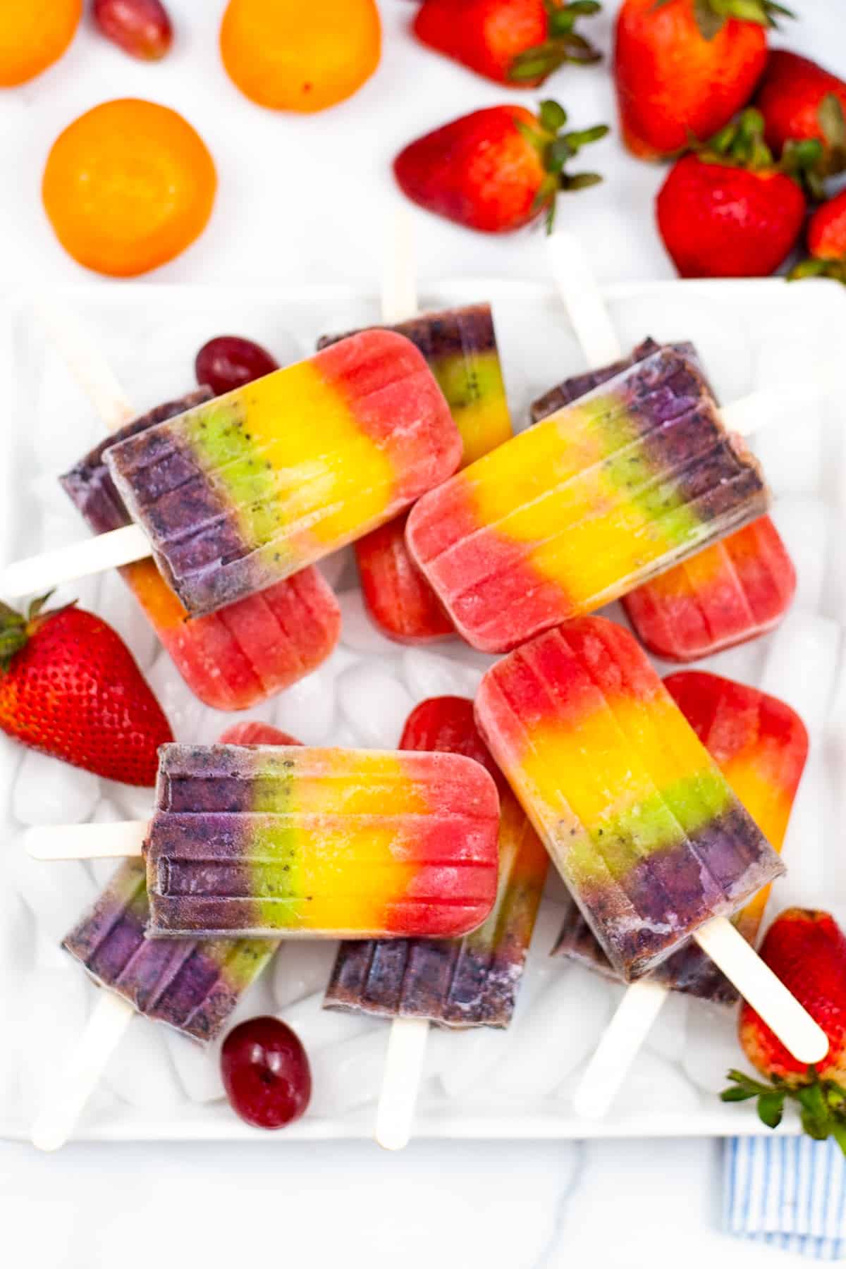 Bright and colorful rainbow fruit popsicles served on a bed of ice on a white plate. Mandarin oranges, strawberries, and grapes surround the plate.