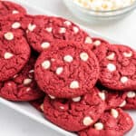 Red velvet cookies with white chocolate chips piled on white serving platter with bowl of white chocolate chips in the background