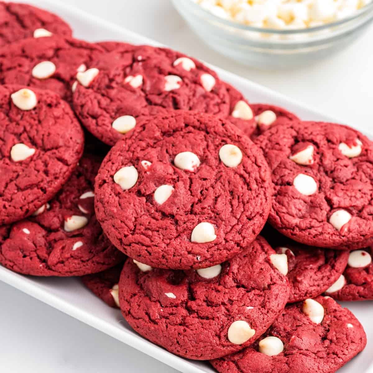 Red velvet cookies with white chocolate chips piled on white serving platter with bowl of white chocolate chips in the background