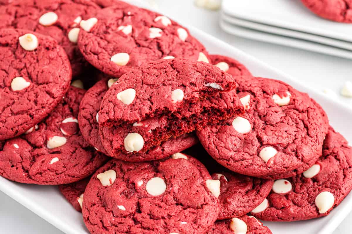 Red velvet cookies with white chocolate chips on white plate. Top cookie is broken in half to show soft and chewy texture. A bowl of white chocolate chips in the background