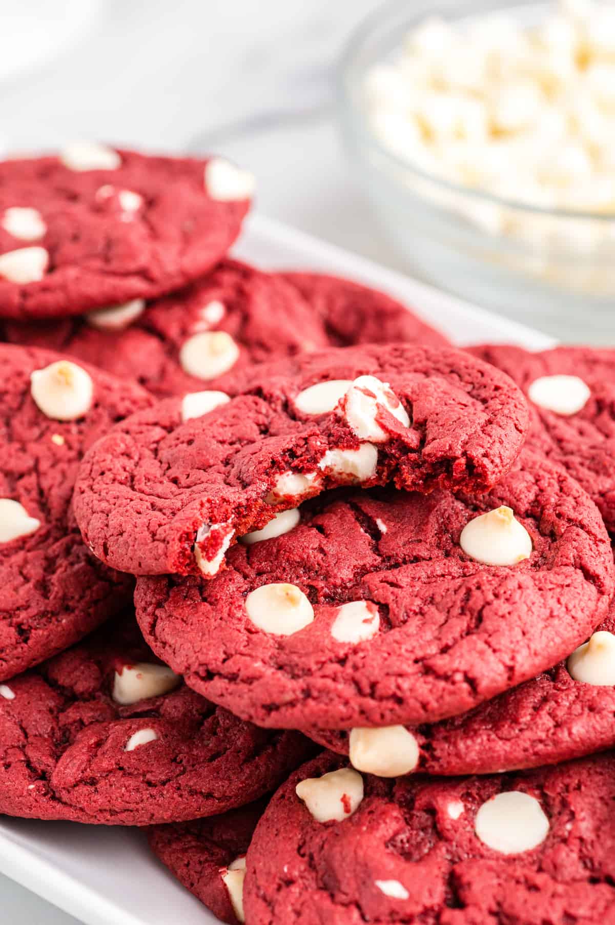 Red velvet cookies with white chocolate chips on white plate. Top cookie has bite taken out of it to show soft and chewy texture. A bowl of white chocolate chips in the background
