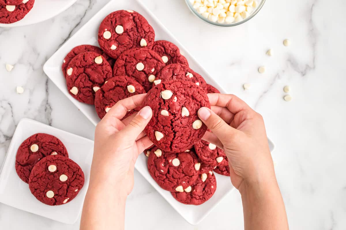 Red velvet cake mix cookies on white platter on countertop. Two hands are reaching into frame and breaking apart one cookie.