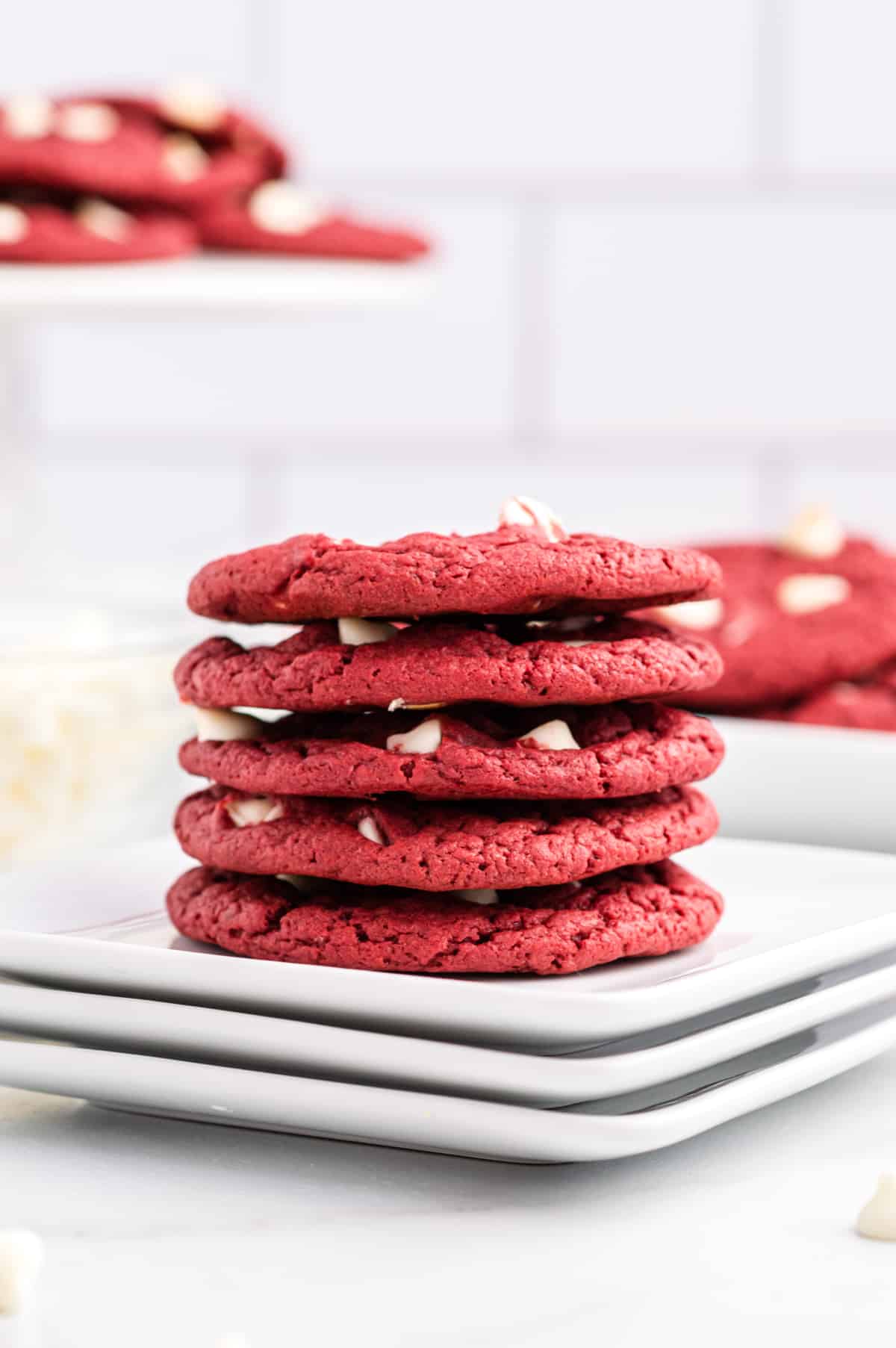 Red velvet white chocolate cookies stacked on white plates. More cookies can be seen in background.