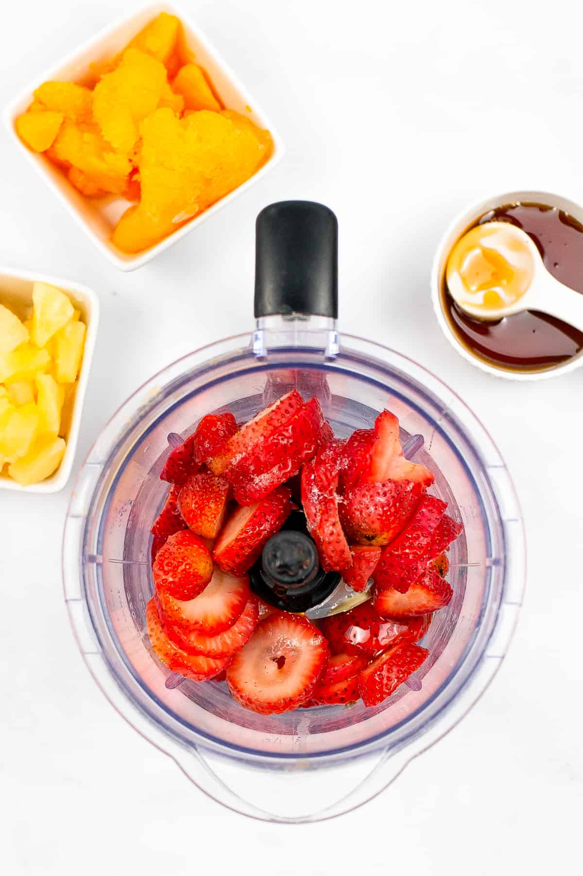 Sliced strawberries in blender with bowls of pineapple chunks, mandarin orange slices, and honey around it