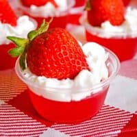 Strawberry jello shots with whipped cream and strawberry on top, served in plastic shot glasses.