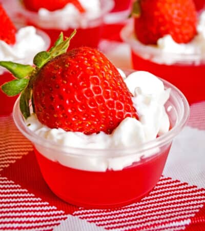Strawberry jello shots with whipped cream and strawberry on top, served in plastic shot glasses.