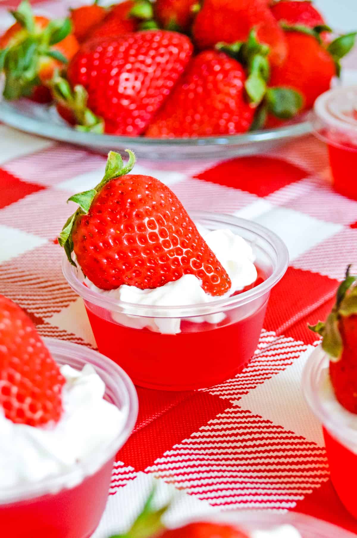 strawberry jello shots on a red checked tablecloth with a plate of fresh strawberries in the background