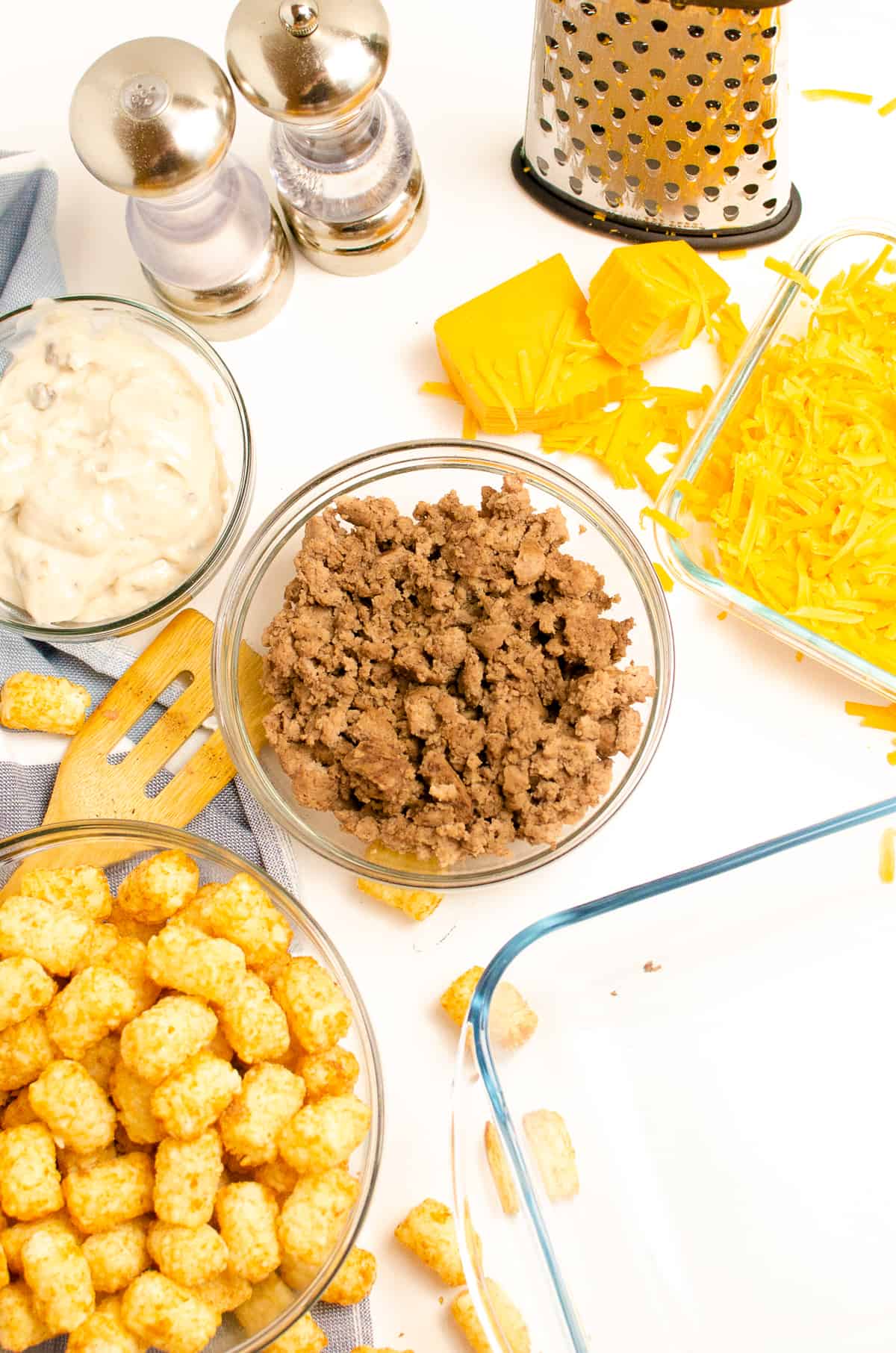 Ingredients in glass bowls: browned ground beef, cream of mushroom soup, cheddar cheese and cheese shredder, frozen tater tots, salt and pepper, and glass casserole dish