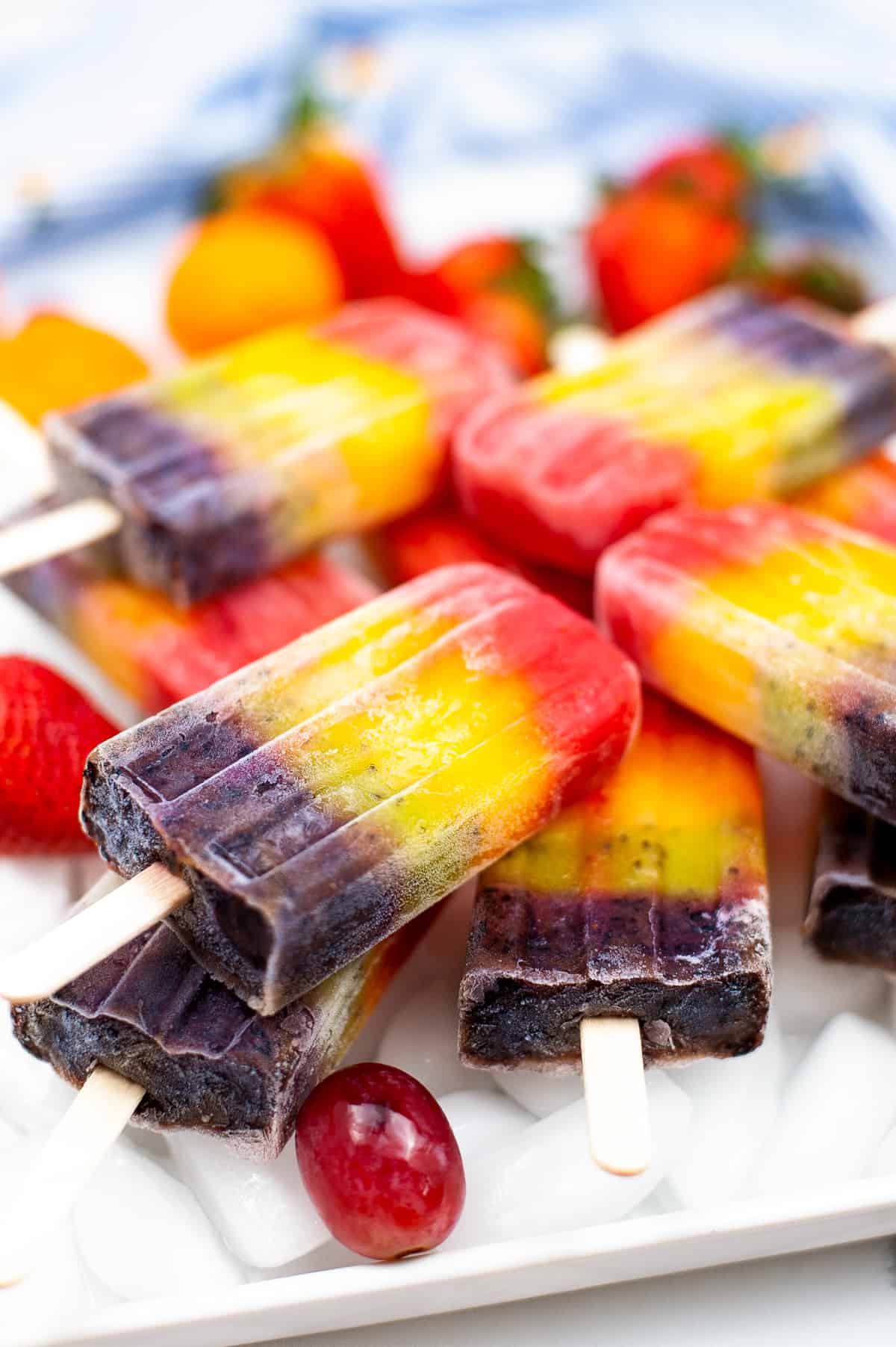 Bright and colorful rainbow fruit popsicles served on a bed of ice on a white plate. Mandarin oranges, strawberries, and grapes surround the plate.