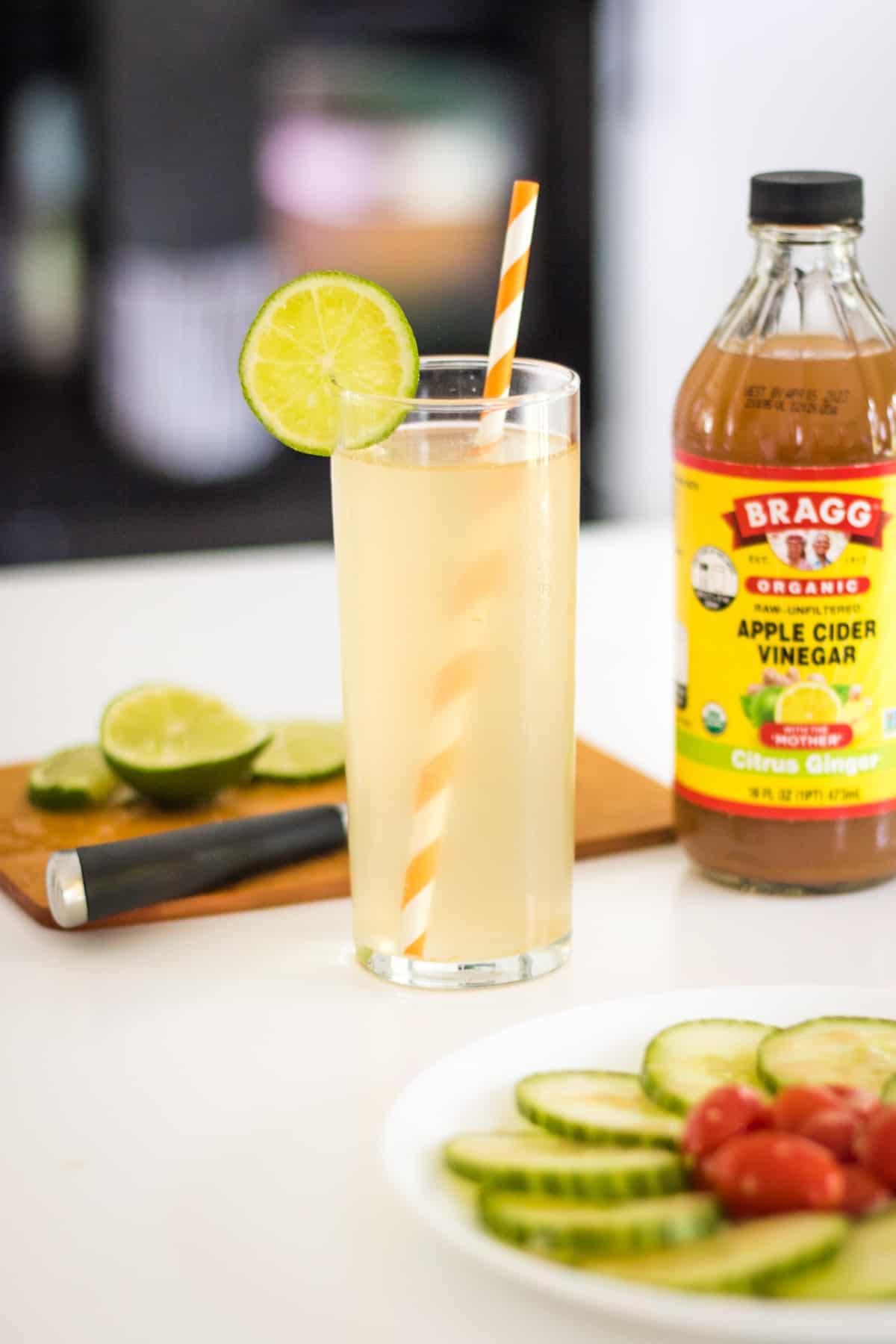 Apple cider vinegar sparkling mocktail garnished with lime with lime on a cutting board and a bottle of apple cider vinegar in the background