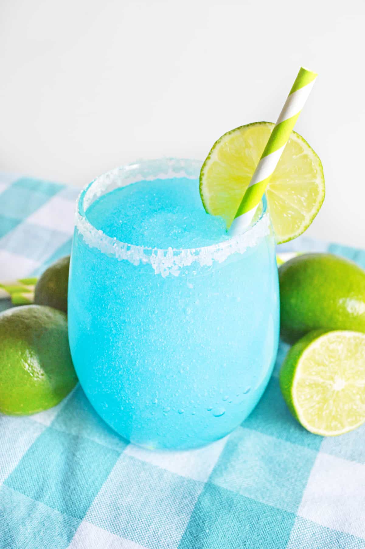 Bright blue frozen margarita garnished with lime and served in a glass rimmed with coarse sugar. Limes are next to glass on blue tablecloth.