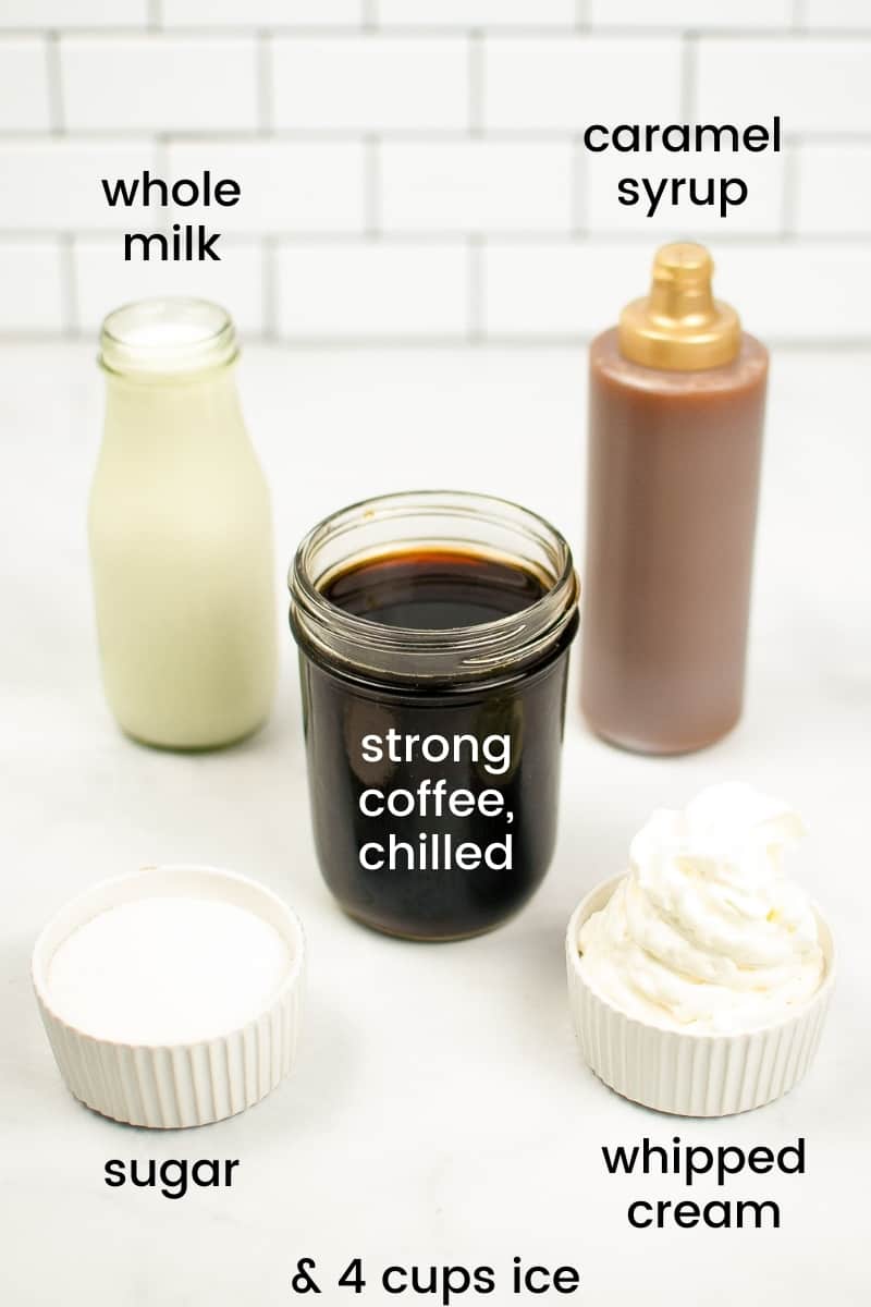 glass jar of whole milk, bottle of caramel syrup, jar of chilled strong coffee, bowl of granulated sugar, and bowl of whipped cream