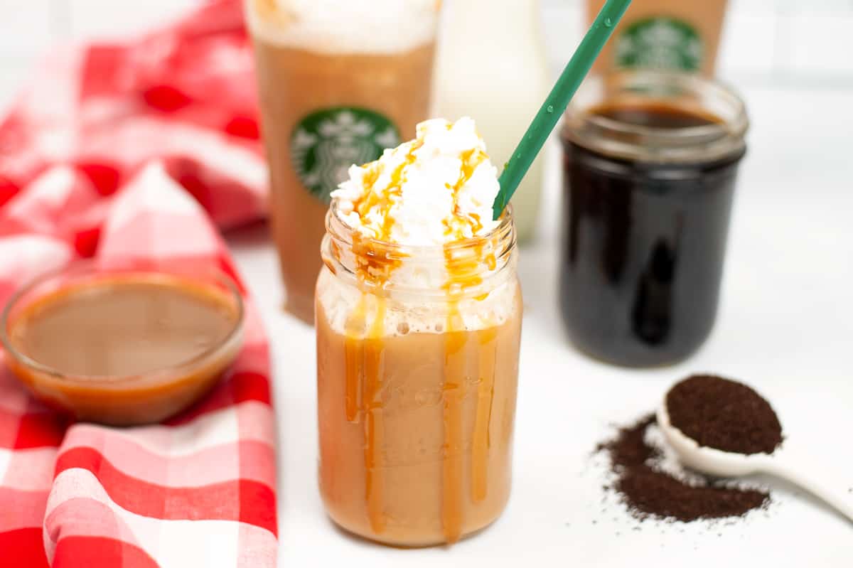 Caramel frappuccino with whipped cream and green straw. Additional cups and a bowl of caramel syrup, cold coffee, and coffee grinds are in background.