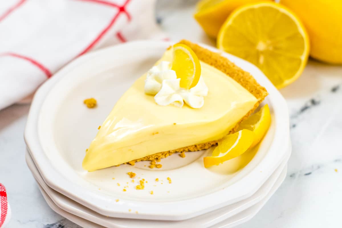 Lemon Icebox Pie slice on white plate, topped with whipped cream and lemon slice