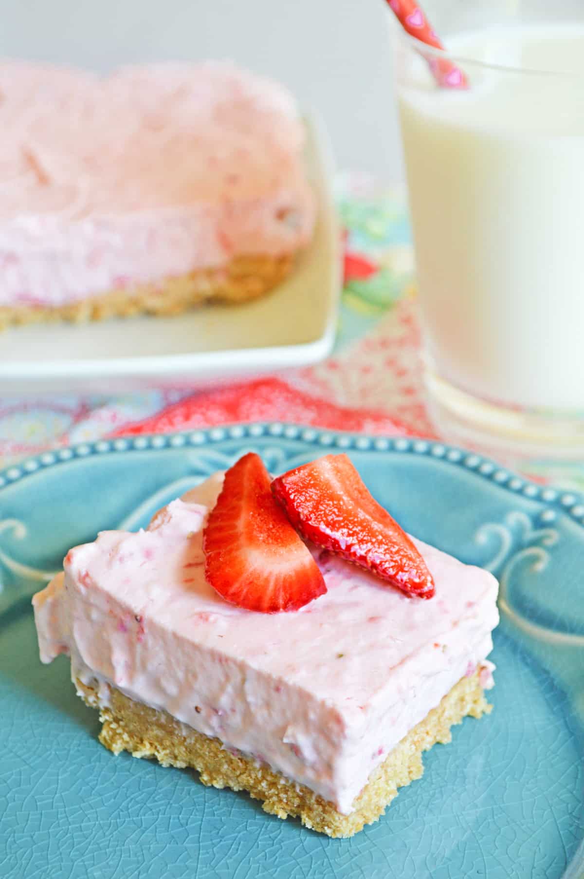 No-Bake Strawberry Cheesecake Bar with a thick graham cracker crust on blue plate topped with fresh strawberries. Glass of milk and remaining bars are in background.