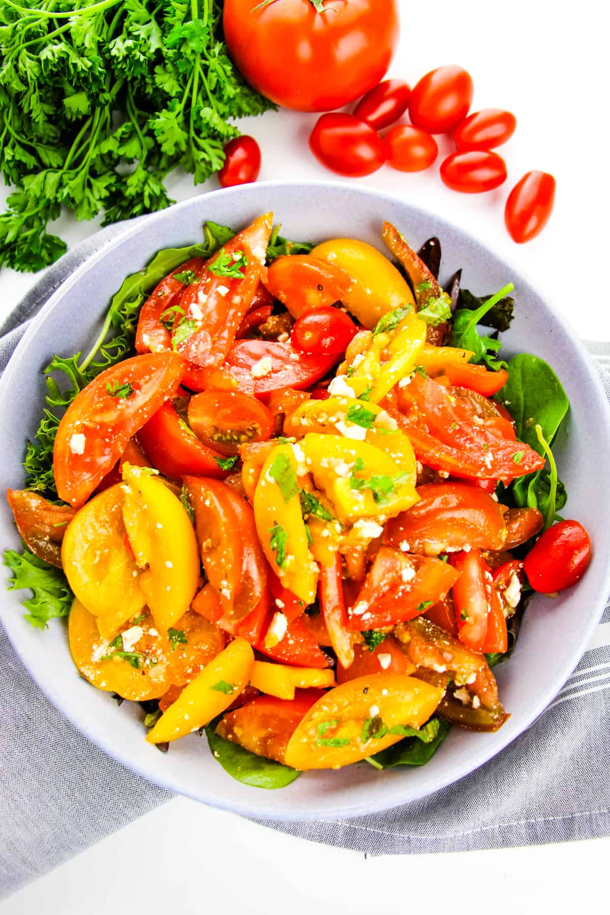 Overhead image of tomato salad in large bowl served over bed of greens. Fresh herbs and tomatoes are in background