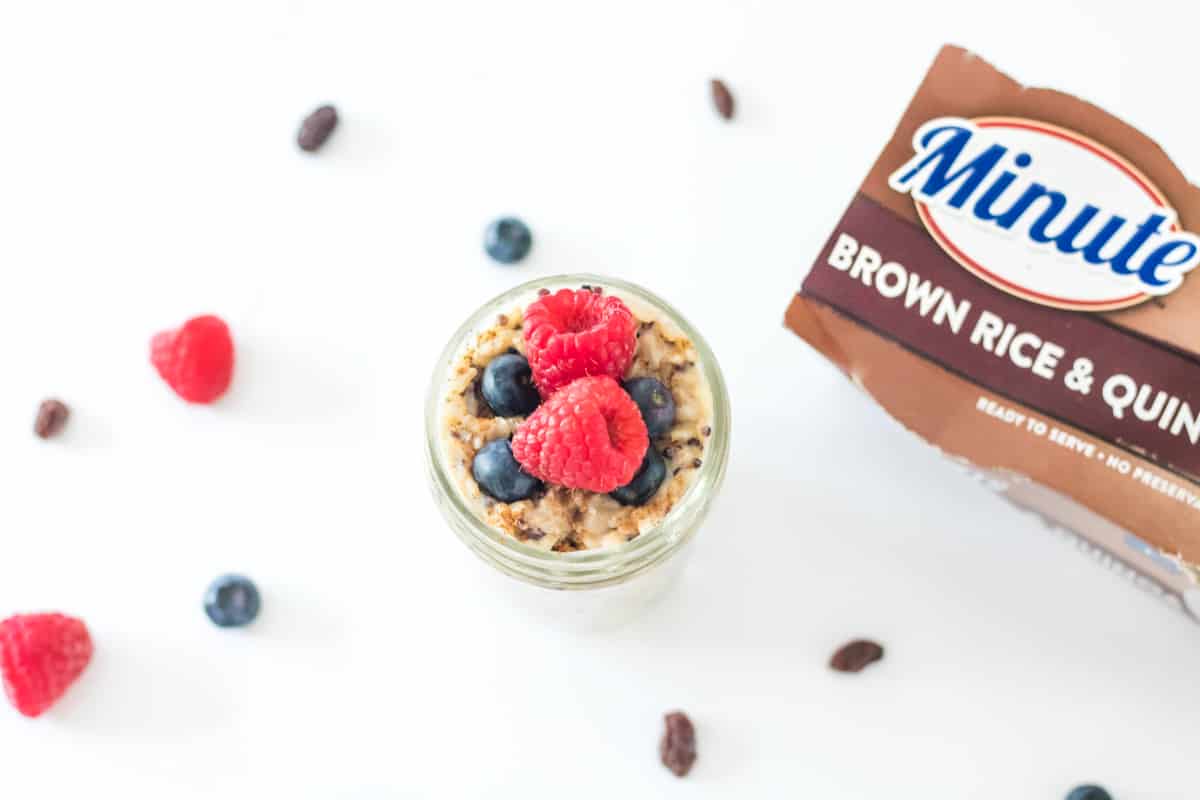 Top-down view of jar of brown rice pudding topped with raspberries and blueberries. Raisins and additional berries are sprinkled on countertop and package of Brown Rice Minute Rice is next to the jar