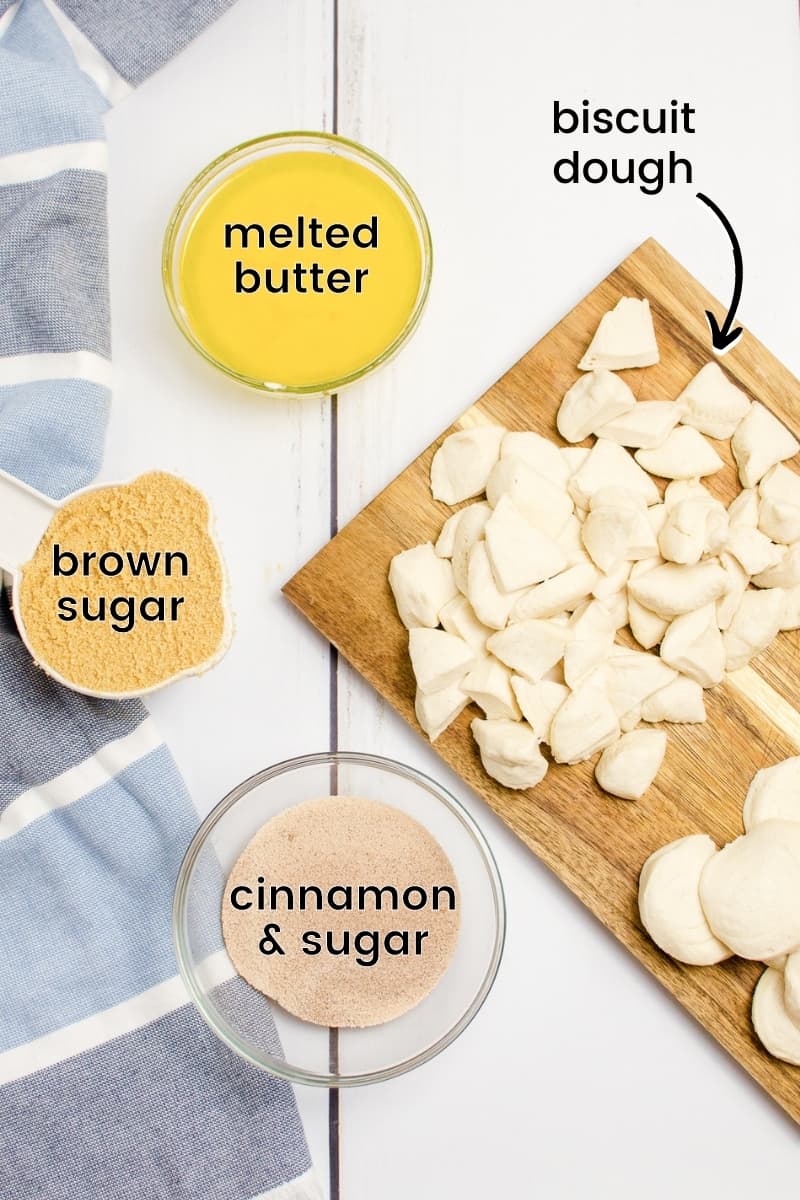 Cutting board with biscuits cut into quarters, bowl of melted butter, bowl of cinnamon and white sugar mixed, and measuring cup of packed brown sugar.
