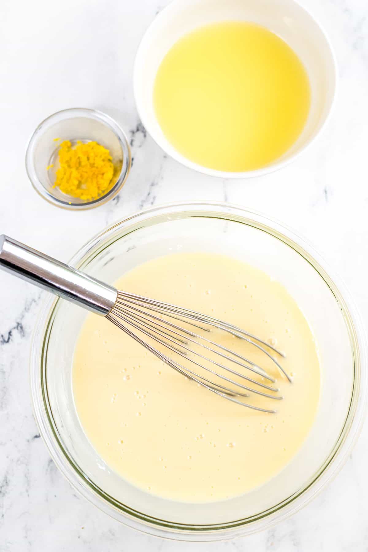 Whisk in bowl of sweetened condensed milk. Lemon zest and lemon juice in bowls next to large mixing bowl.