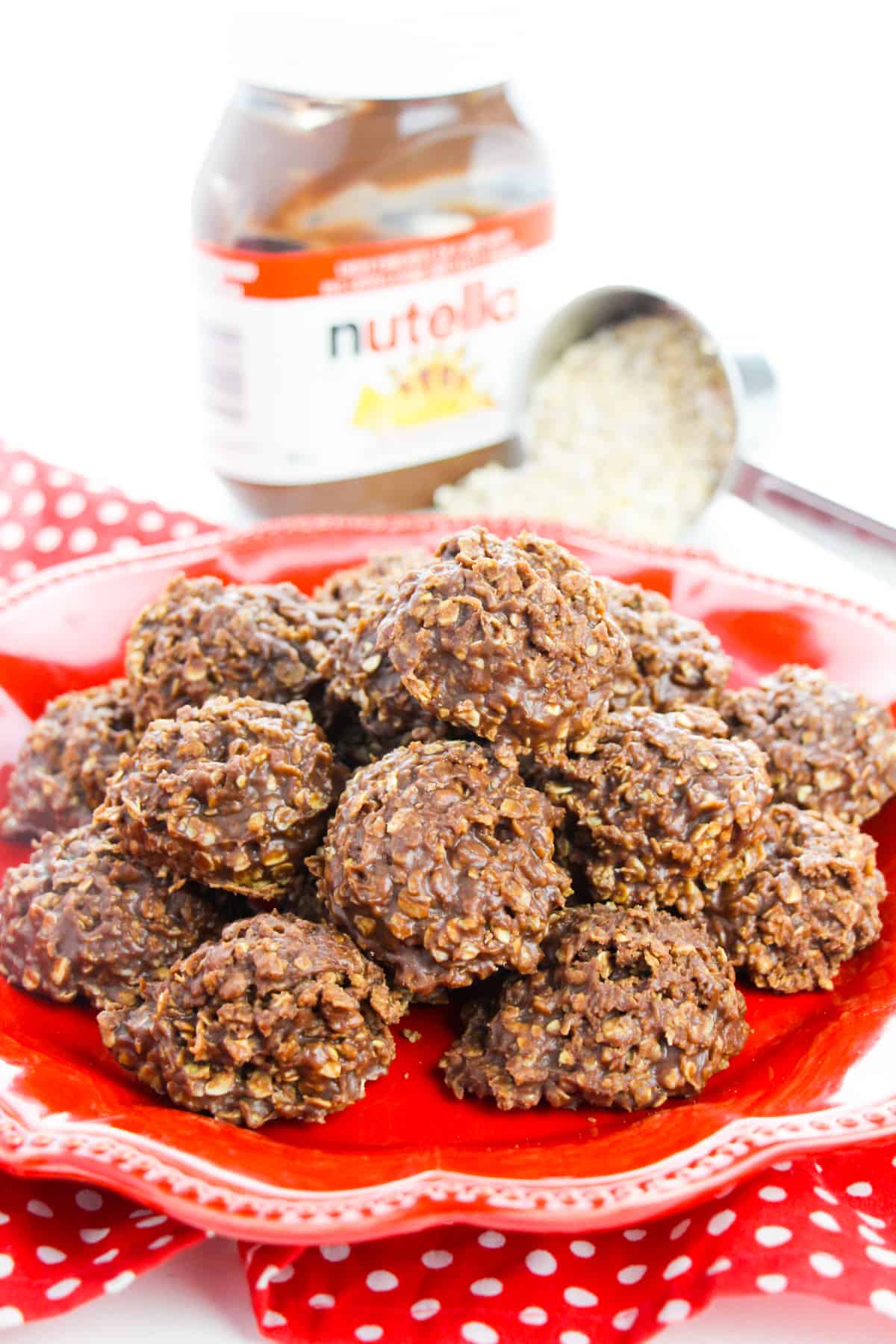 No-bake Nutella Cookies piled on a red plate with scoop of oats and jar of nutella spread behind them