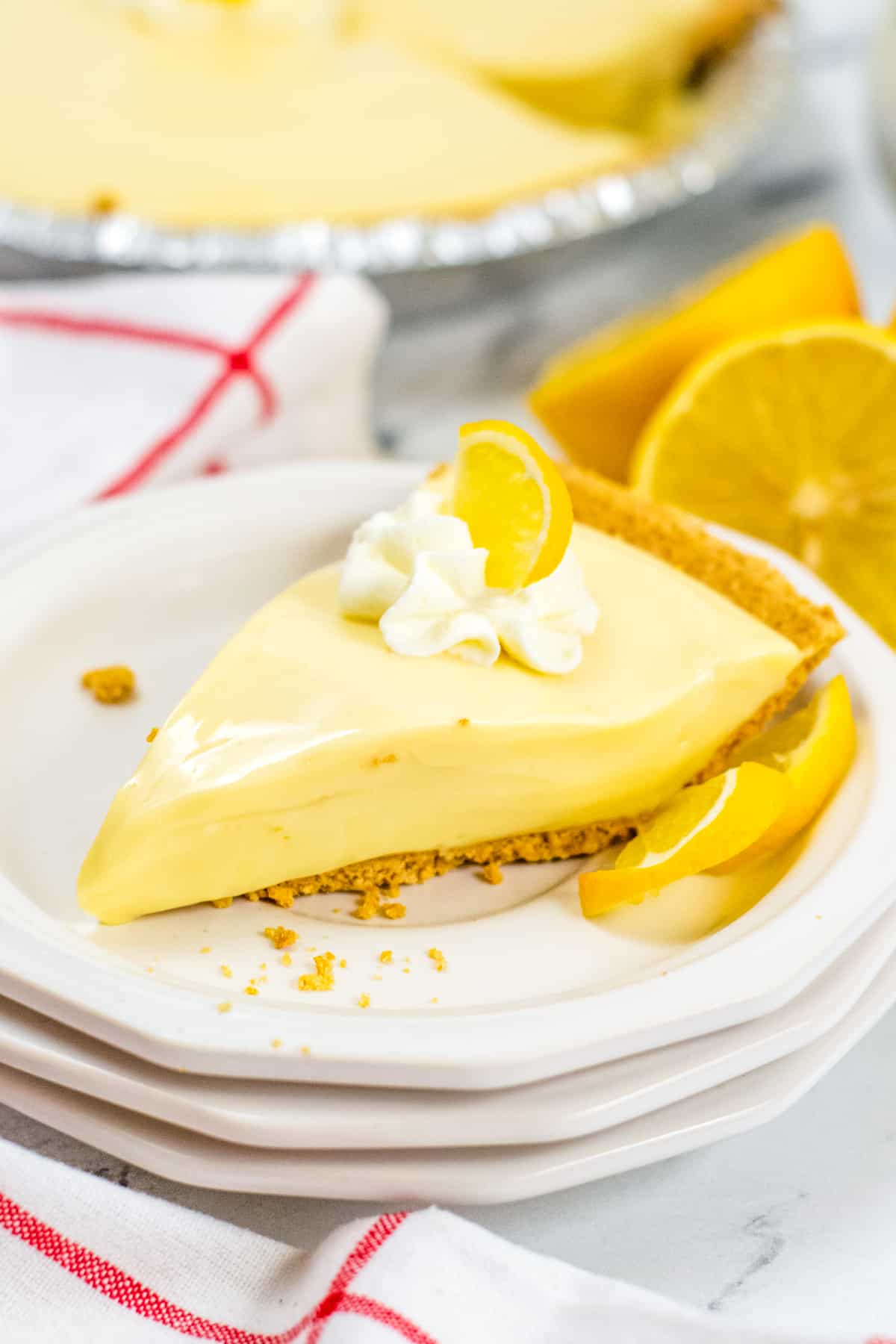 Lemon Icebox Pie slice on stack of white plates. Pie is topped with dollop of whipped cream and slice of lemon. Whole pie can be seen in background, as well as fresh lemons