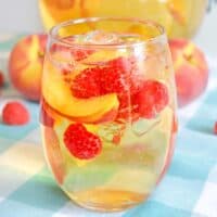 White peach sangria with fresh peaches and raspberries in a stemless wine glass with whole peaches in background