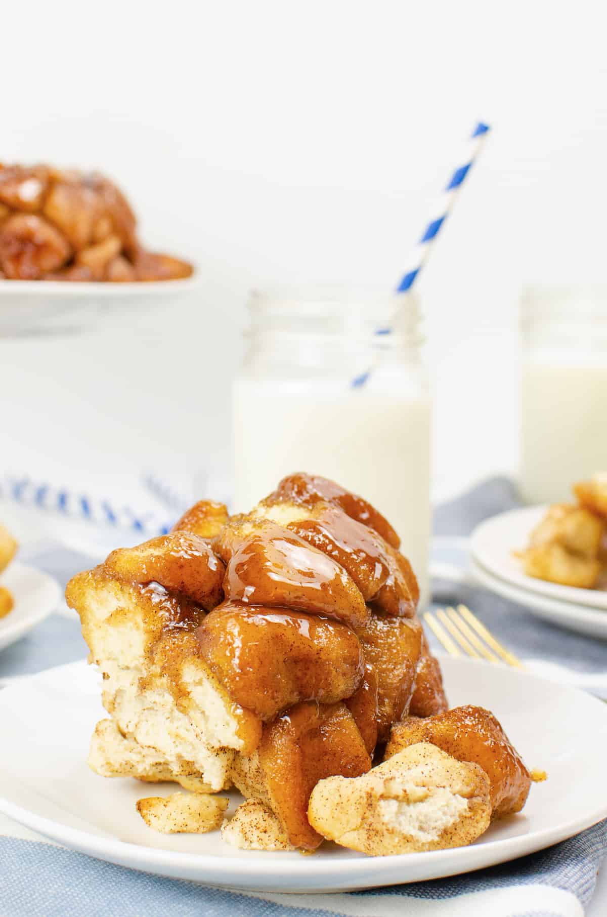 Cinnamon sugar monkey bread serving on white plate with whole monkey bread and glass of milk with straw in background.