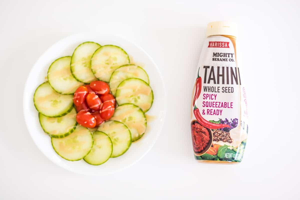 Cucumbers slices and cherry tomatoes drizzles with tahini. Bottle of Mighty Sesame whole seed spicy squeezable and ready next to the salad