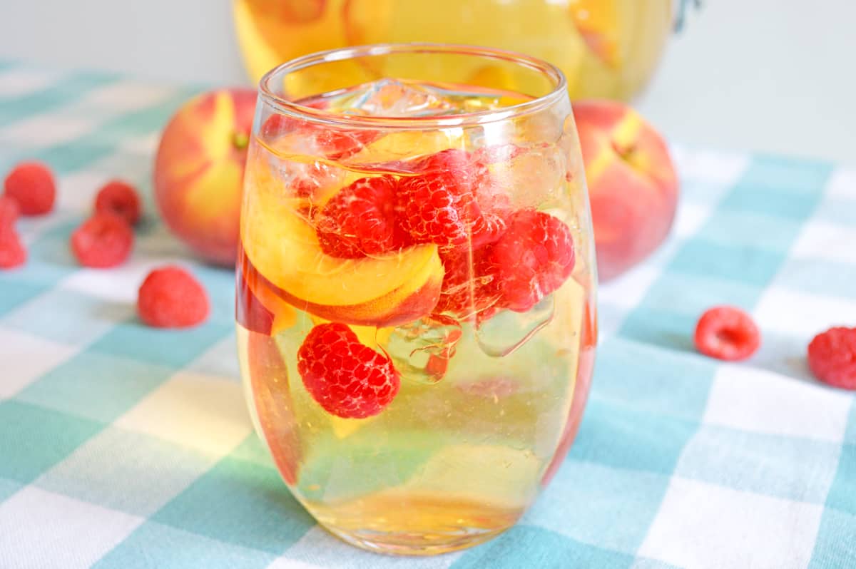 White peach sangria with fresh peaches and raspberries in a stemless wine glass with whole peaches and raspberries in background