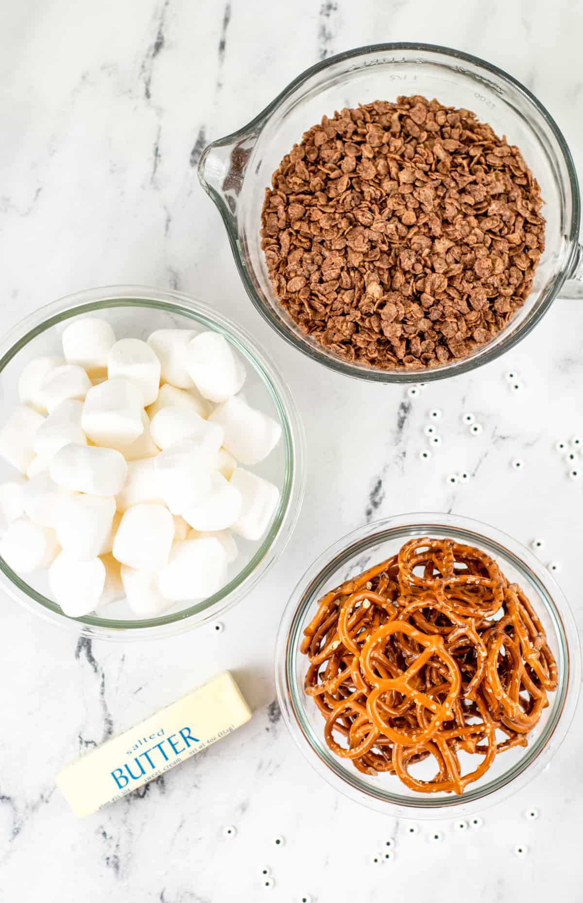 Spider Krisipies Treat Ingredients on countertop: bowl of marshmallows, bowl of chocolate krispy cereal, stick of butter, bowl of thin pretzels