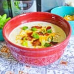 crockpot loaded baked potato soup in red bowl topped with bacon, scallions, and cheese.