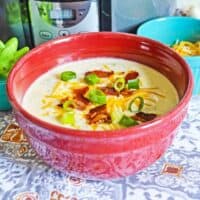 crockpot loaded baked potato soup in red bowl topped with bacon, scallions, and cheese.