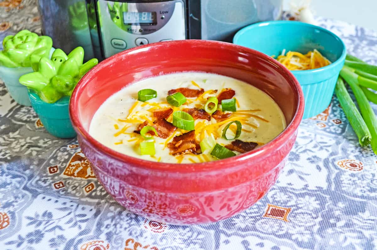 bowl of loaded backed potato soup topped with bacon, scallions, and cheese. Crockpot, scallions, and shredded cheese are in the background.