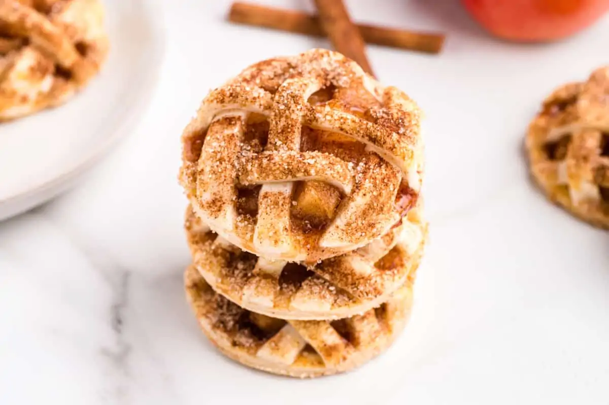 Apple pie cookies with lattice top stacked on top of one another. More cookies and a few cinnamon sticks are in background.