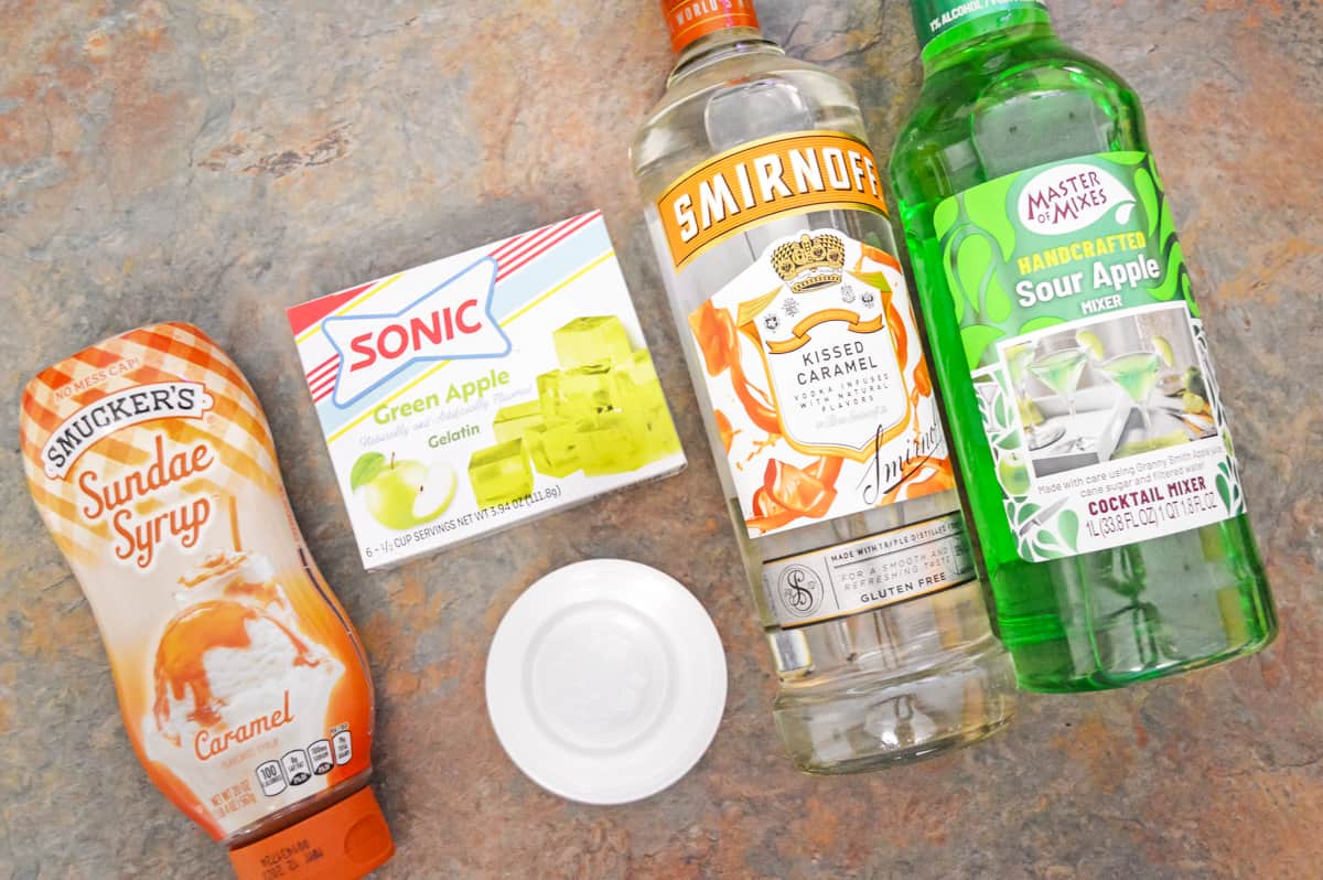 On countertop: Bottle of Smucker's Caramel Sundae Syrup, box of Sonic Green Apple Jello, bottle of Smirnoff Kissed Caramel Vodka, bottle of Master Mixes sour apple mixer, and small bowl of water
