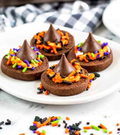 Four chocolate witch hat cookies with Hershey kisses on a white plate with halloween sprinkles around the table.