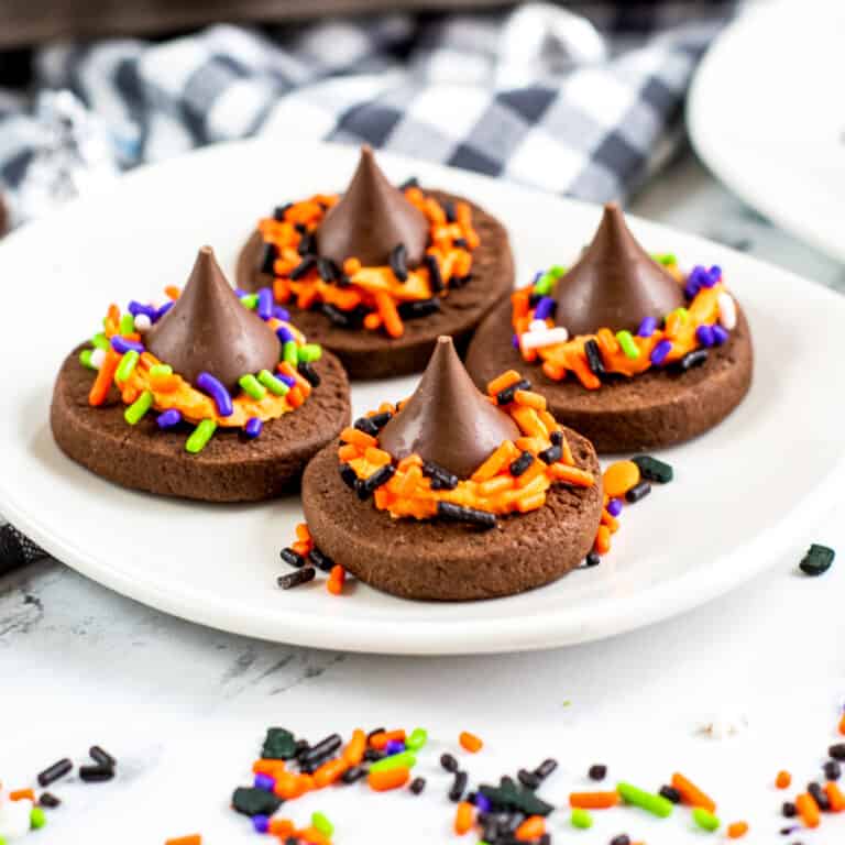 Four chocolate witch hat cookies with Hershey kisses on a white plate with halloween sprinkles around the table.